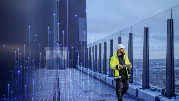 Homem com colete de alta visibilidade e capacete caminhando pelo corredor de um edifício comercial com um tablet na mão